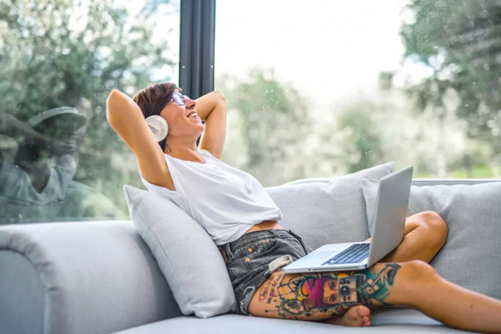 Woman reclining on a couch, using laptop while listening to music with headphones, smiling and relaxed.